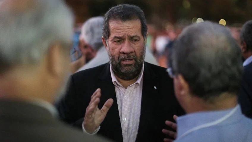 Um homem com barba e vestindo um terno, possivelmente o ministro da Previdência, fala animadamente em meio a uma reunião ao ar livre. O fundo está desfocado, sugerindo uma multidão discutindo políticas governamentais.